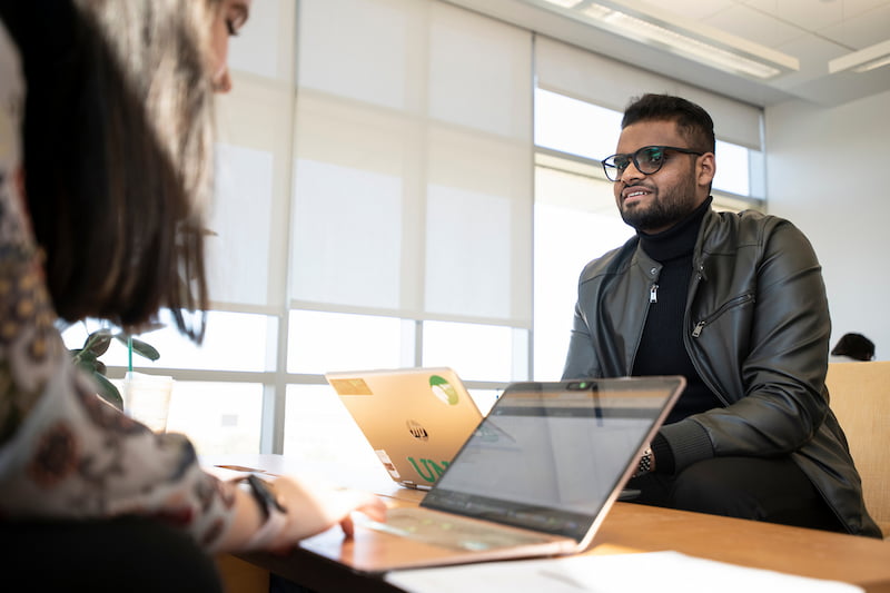 A man wearing glasses and a black leather jacket is sitting at a table with laptops in front of him. A woman is partially visible in the foreground, also at the table.