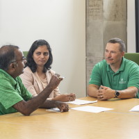 Three people are sitting at a table and appear to be in a meeting. One man is gesturing with his hand as he speaks to the other two.