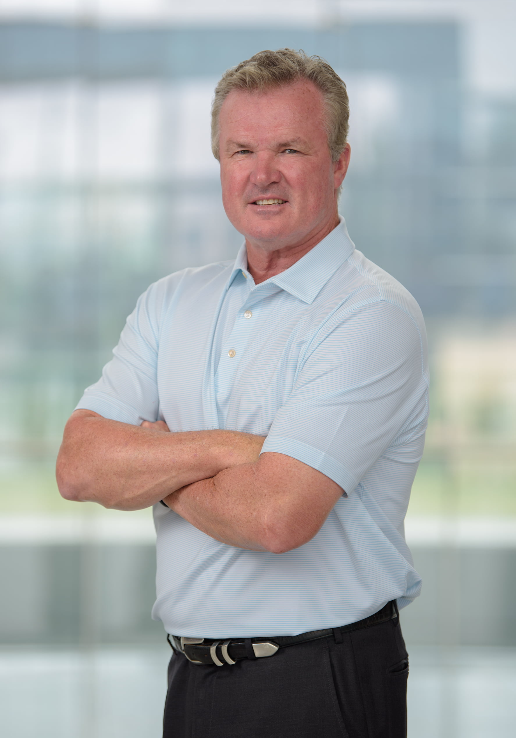 A middle-aged man with graying hair and a light blue striped polo shirt stands with his arms crossed. He is looking at the camera with a slight frown. The background is blurred and appears to be an office building interior with large windows.