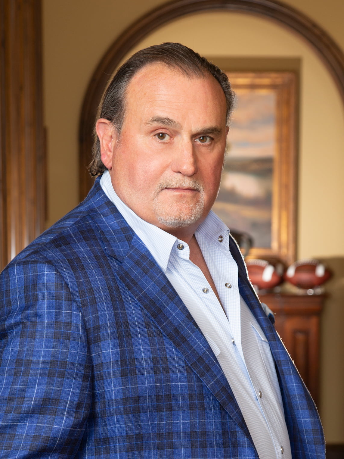 A middle-aged man in a blue plaid suit jacket and a light blue collared shirt looks directly at the camera. In the background, a painting and three footballs are visible on a wooden surface.