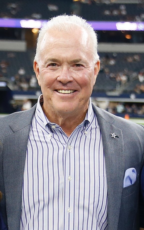 A man with white hair and a blue and white striped shirt smiles at the camera. He is wearing a gray pinstripe suit jacket with a blue pocket square and a star pin on the lapel.