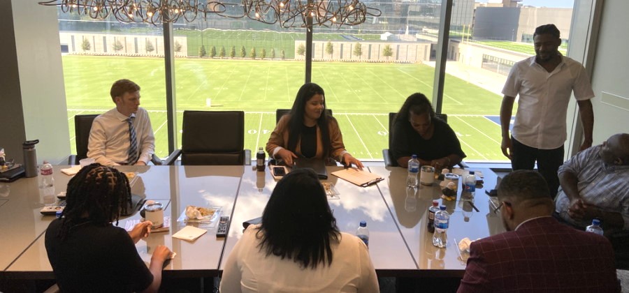 A diverse group of people are gathered around a conference table in a room overlooking a football field. Some are seated, engaged in discussion, while one man stands.