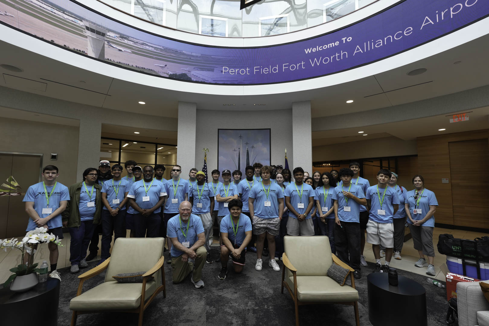 A large group of young people wearing matching blue t-shirts with "ACE CAMP" printed on them are posing for a group photo. They are standing in an airport with a large image displaying an airport scene and the words "Welcome To Perot Field Fort Worth Alliance Airport."