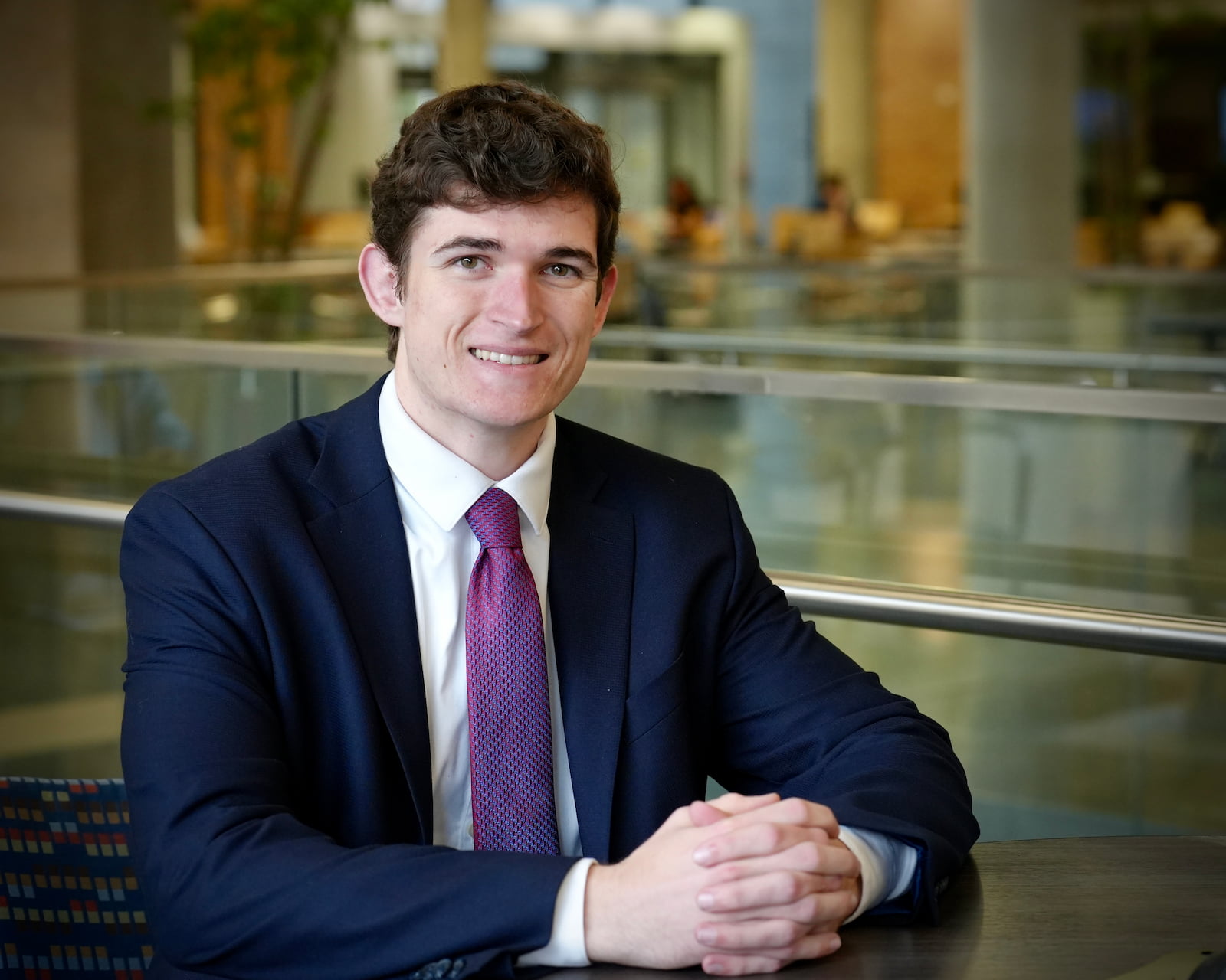 A young man in a suit and tie smiles while sitting with his hands clasped at a table. The background is blurred with people and plants visible.