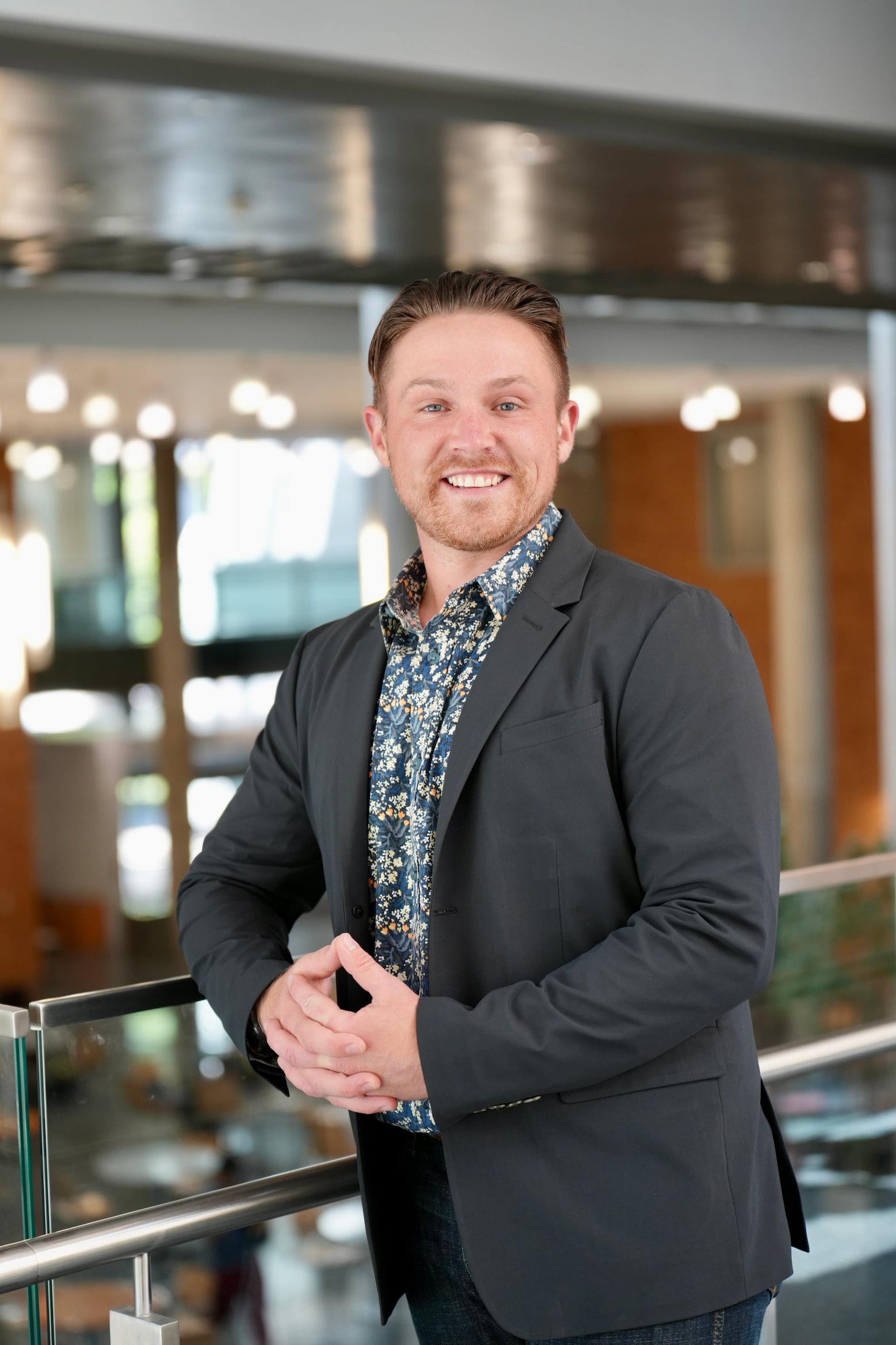 A young, smiling man in a dark suit jacket and a floral shirt leans on a railing indoors. He has his hands clasped in front of him.