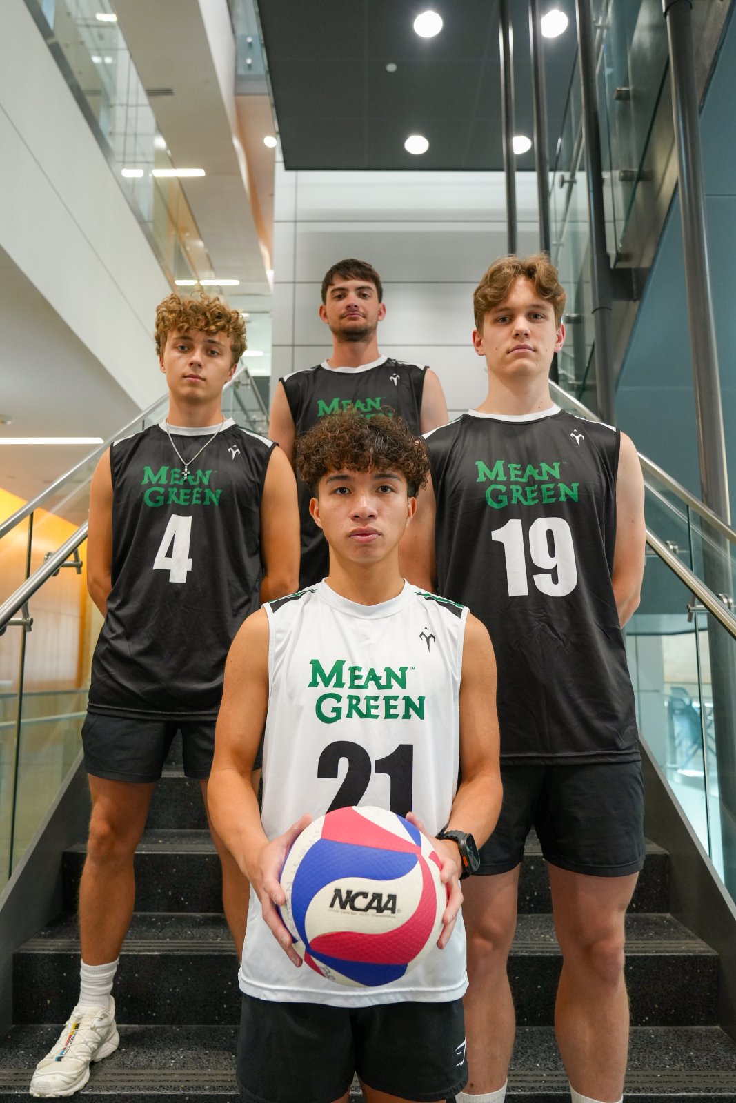 Four young men in volleyball uniforms stand on a staircase, posing for a photo with a volleyball. They are wearing sleeveless jerseys with "Mean Green" printed on them and their respective numbers.