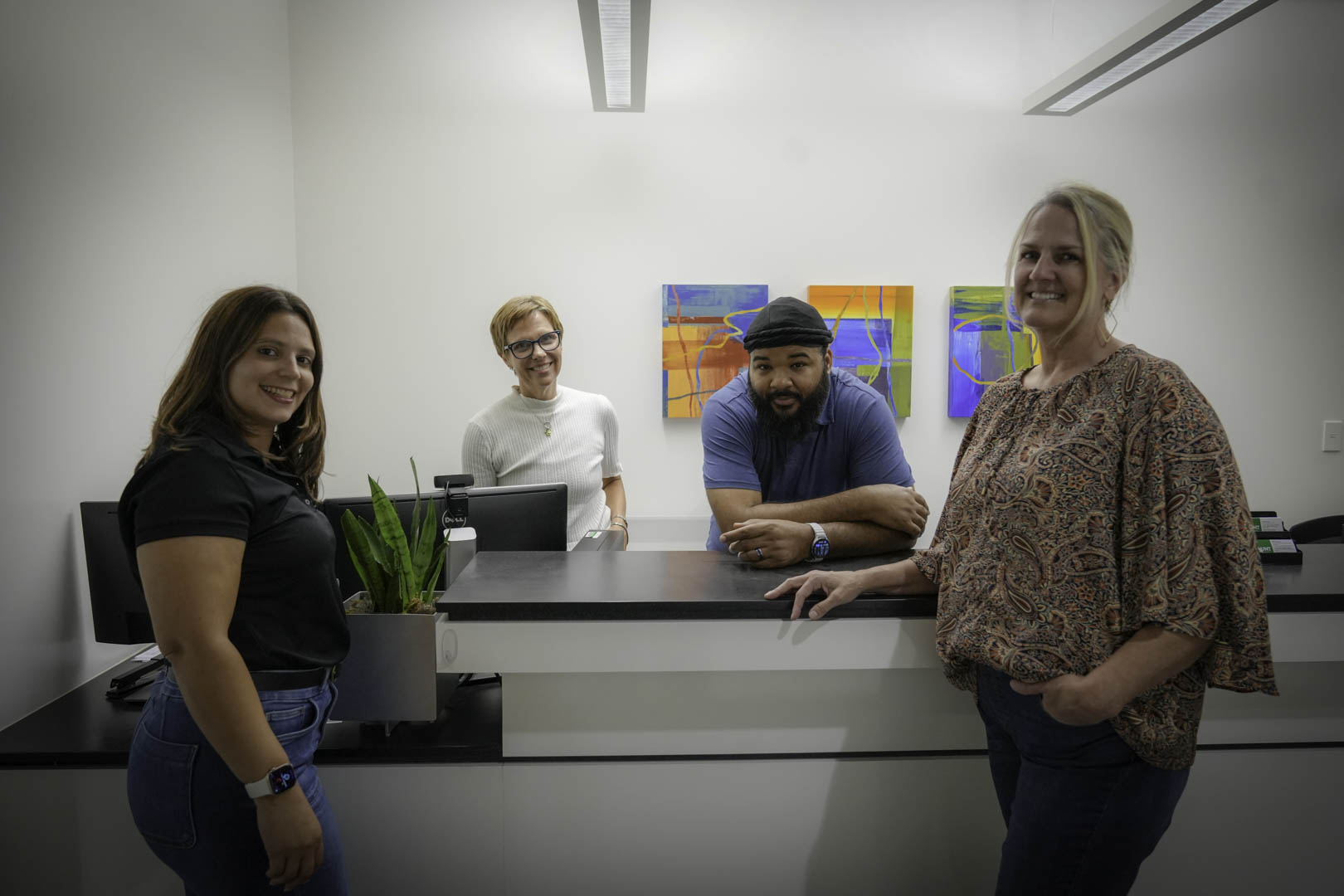 Four individuals stand behind a reception desk in an office. They are all smiling and looking towards the camera. The background features modern abstract art.