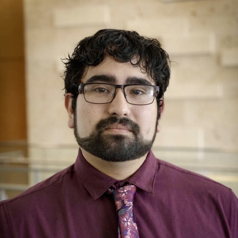 A close-up portrait of Ruben Garcia wearing glasses and a purple shirt with a floral tie. He has dark, curly hair and a beard.