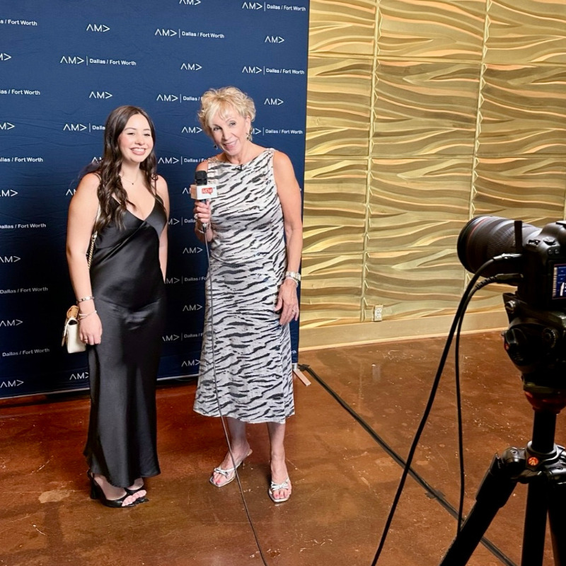 Shelby King, wearing a black dress, is being interviewed by an older woman in a zebra-print dress in front of a blue backdrop with the American Marketing Association - Dallas/Ft. Worth logo on it. A camera on a tripod is recording them.