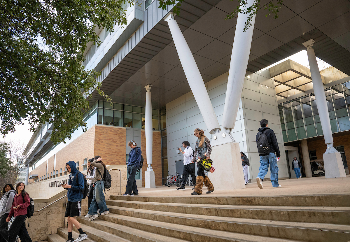 Students are walking up and down the wide concrete stairs in front of a modern building. The building has large white support beams and a brick facade.